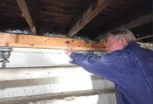 A man in coveralls standing in a crawlspace beneath a home points to a board on top of a concrete foundation